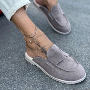 May include: A pair of grey suede slip-on loafers featuring a rounded toe, a wide strap across the vamp, and white stitching details. The shoes have a flat sole and an open back design, providing a casual look with a soft, textured finish.