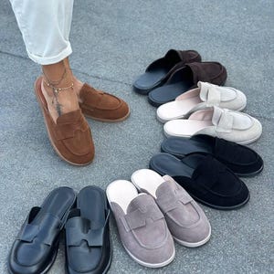 May include: A collection of slip-on loafers in various colors including brown, black, grey, and beige. Each shoe features a rounded toe, a flat sole, and a distinctive cutout detail on the upper strap. The loafers are arranged in a semi-circle.