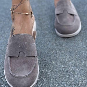 May include: A pair of taupe suede slip-on loafers featuring a rounded toe, a wide strap across the vamp with a central cutout detail, and white contrast stitching along the edges. The shoes have a flat, white rubber sole and a low-cut heel design.