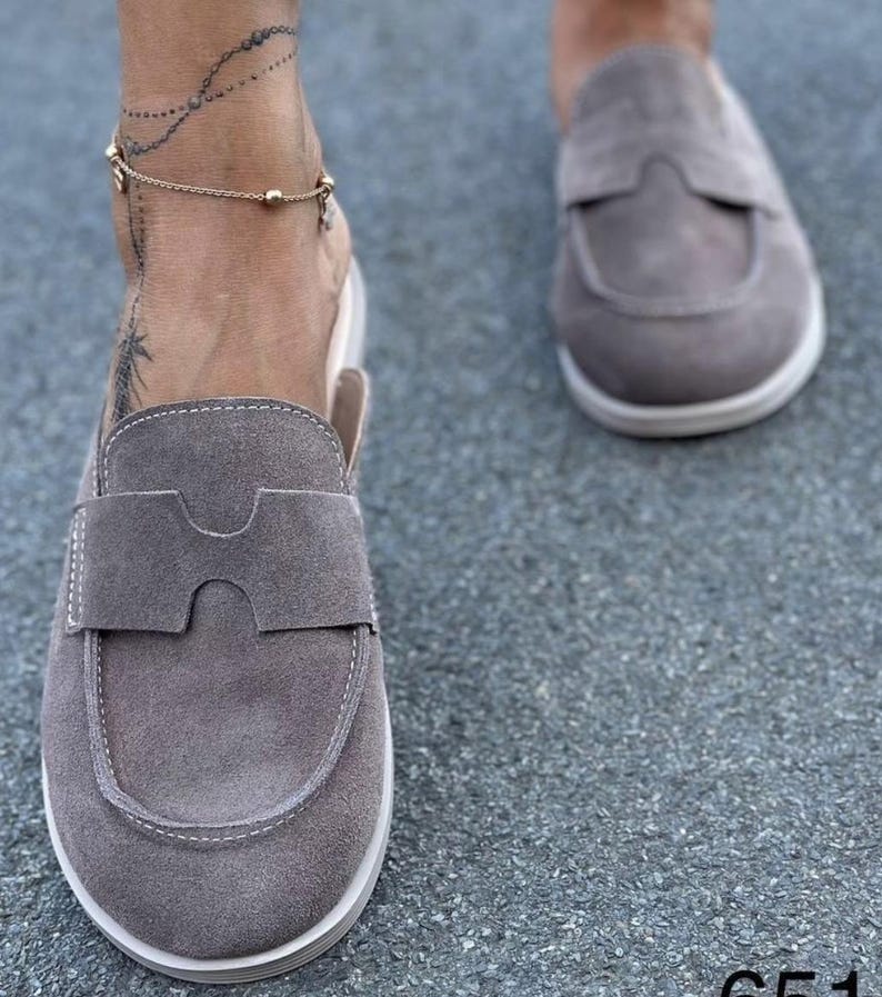 May include: A pair of taupe suede slip-on loafers featuring a rounded toe, a wide strap across the vamp with a central cutout detail, and white contrast stitching along the edges. The shoes have a flat, white rubber sole and a low-cut heel design.