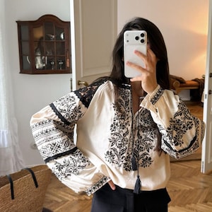 Puede incluir: Una blusa de manga larga color crema con intrincados bordados florales negros en el pecho y las mangas, acentuada con inserciones de encaje negro en los hombros y brazos, y un lazo en el cuello terminado con dos borlas negras.