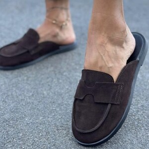 May include: A pair of dark brown suede backless loafers featuring a rounded toe, a wide strap across the vamp with a cutout detail, and a flat black sole. The slip-on footwear has a soft textured finish and a simple construction for easy wear.