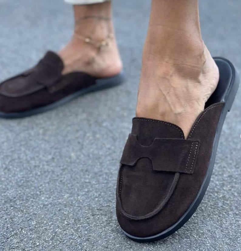 May include: A pair of dark brown suede backless loafers featuring a rounded toe, a wide strap across the vamp with a cutout detail, and a flat black sole. The slip-on footwear has a soft textured finish and a simple construction for easy wear.