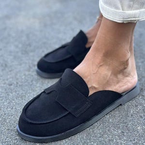 May include: A pair of black suede backless loafers featuring a rounded toe, a decorative cutout strap across the vamp, and a flat, textured rubber sole. The slip-on footwear has a clean, minimalist design with visible stitching along the edges.