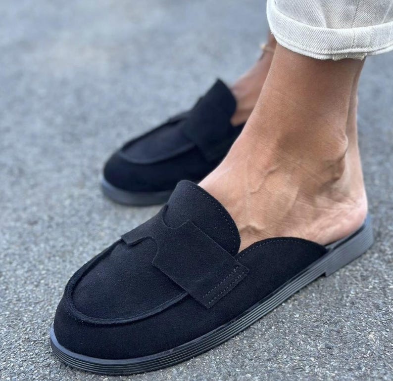 May include: A pair of black suede backless loafers featuring a rounded toe, a decorative cutout strap across the vamp, and a flat, textured rubber sole. The slip-on footwear has a clean, minimalist design with visible stitching along the edges.