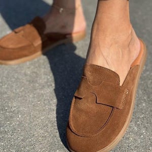 May include: A pair of brown suede mule loafers featuring a rounded toe, a flat sole, and a wide strap across the vamp with a cutout detail. The slip-on footwear has a smooth tan interior lining and a textured brown outsole for traction.