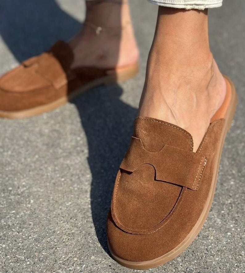 May include: A pair of brown suede mule loafers featuring a rounded toe, a flat sole, and a wide strap across the vamp with a cutout detail. The slip-on footwear has a smooth tan interior lining and a textured brown outsole for traction.