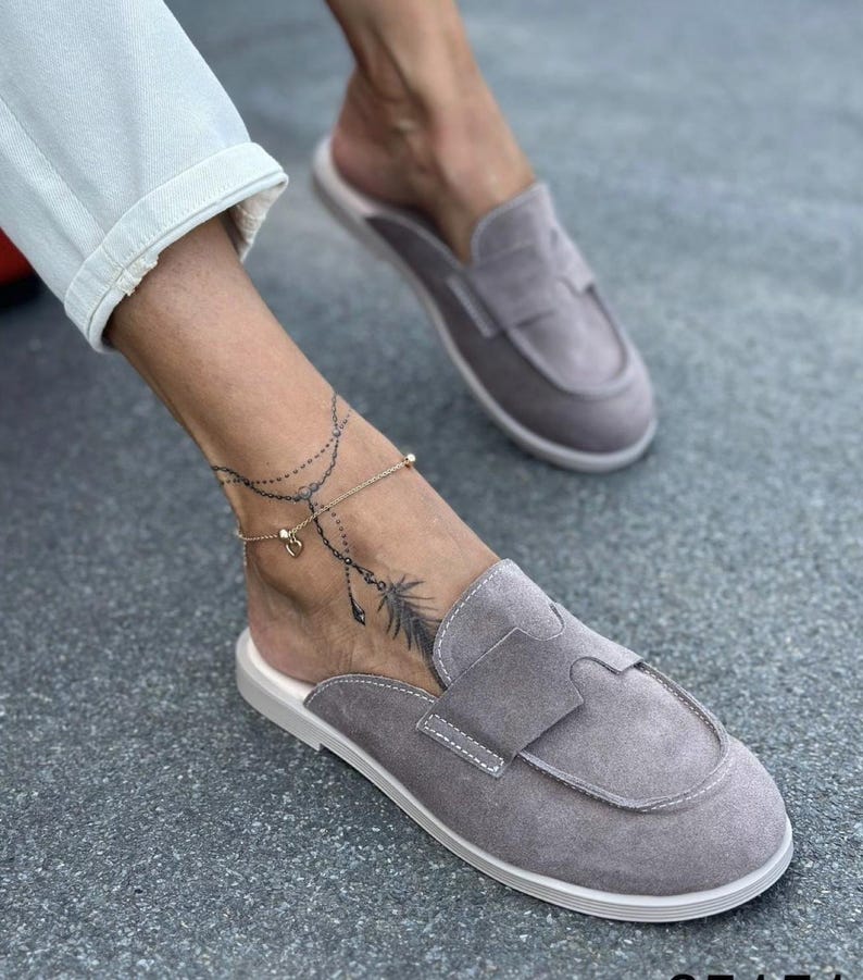 May include: A pair of grey suede slip-on loafers featuring a rounded toe, a wide strap across the vamp, and white stitching details. The shoes have a flat sole and an open back design, providing a casual look with a soft, textured finish.
