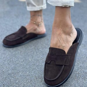May include: A pair of dark brown suede backless loafers featuring a rounded toe, a decorative cutout strap across the vamp, and a flat black sole. The slip-on footwear has a soft textured finish and a clean, simple design for casual wear.