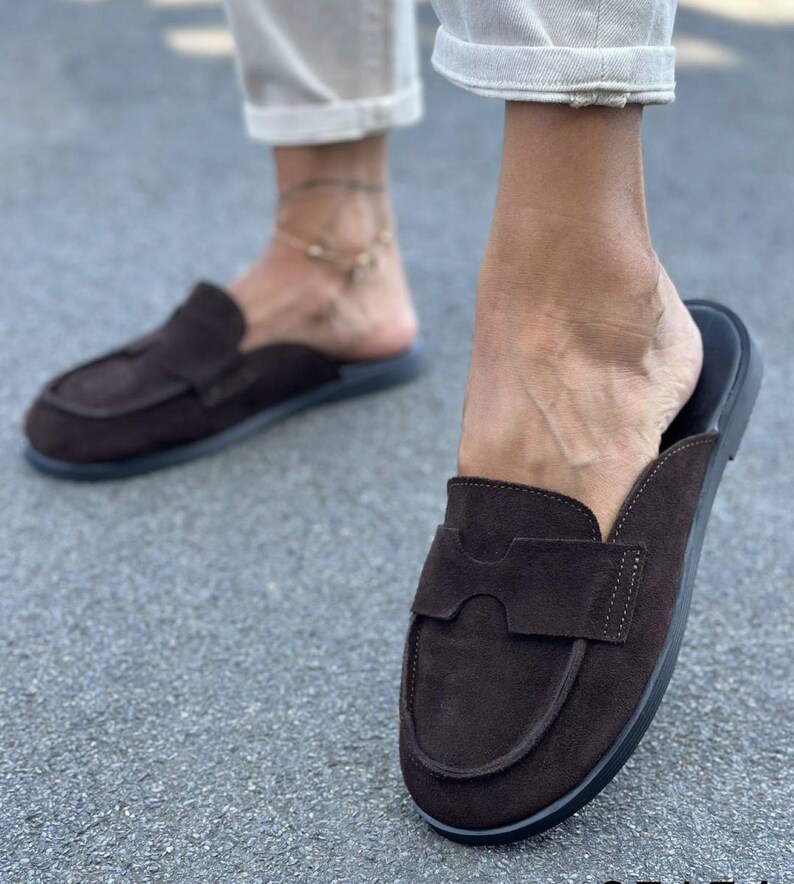 May include: A pair of dark brown suede backless loafers featuring a rounded toe, a decorative cutout strap across the vamp, and a flat black sole. The slip-on footwear has a soft textured finish and a clean, simple design for casual wear.