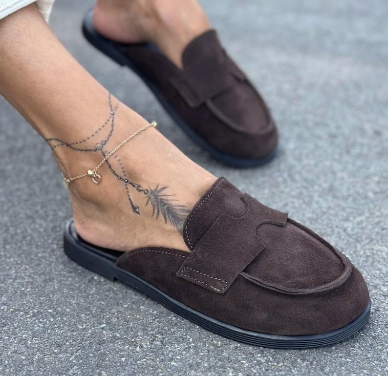 May include: Brown suede slip-on loafers with a black sole. The shoes have a classic design with a strap across the top. The image shows a pair of the shoes, with one shoe in focus.