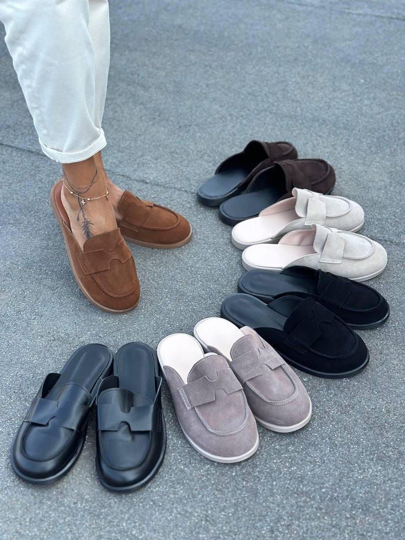 May include: A collection of slip-on loafers in various colors including brown, black, grey, and beige. Each shoe features a rounded toe, a flat sole, and a distinctive cutout detail on the upper strap. The loafers are arranged in a semi-circle.