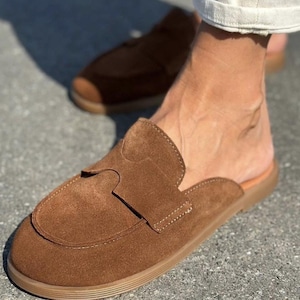 May include: A pair of brown suede slip-on loafers featuring a rounded toe, a wide strap across the vamp with a cutout detail, and a flat, tan-colored rubber sole. The shoes have a backless design, exposing the heel, and visible stitching.