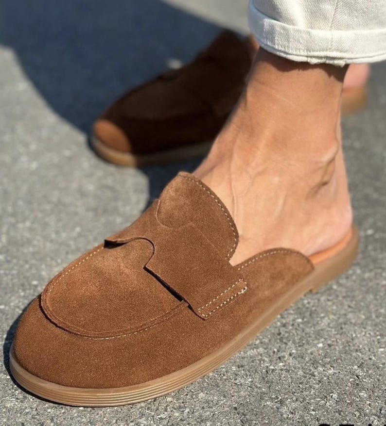 May include: A pair of brown suede slip-on loafers featuring a rounded toe, a wide strap across the vamp with a cutout detail, and a flat, tan-colored rubber sole. The shoes have a backless design, exposing the heel, and visible stitching.
