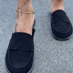 May include: A pair of black suede slip-on loafers featuring a rounded toe and a wide strap across the vamp with a cutout detail. The shoes have a flat sole and a matte finish, providing a simple and functional design for casual footwear.
