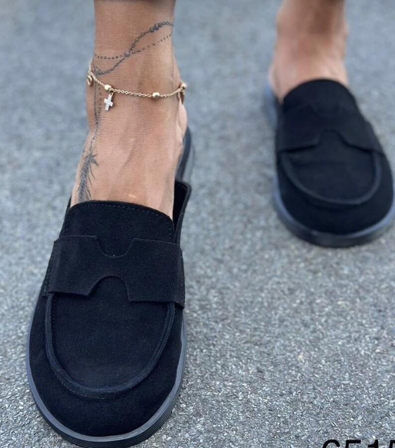 May include: A pair of black suede slip-on loafers featuring a rounded toe and a wide strap across the vamp with a cutout detail. The shoes have a flat sole and a matte finish, providing a simple and functional design for casual footwear.