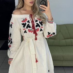 May include: An off-the-shoulder white dress featuring long sleeves and a tie-waist belt. The garment is decorated with black and red floral embroidery patterns, including red tassels at the neckline and striped cuffs on the sleeves.
