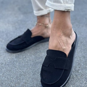 May include: A pair of black suede slip-on loafers featuring a rounded toe, a flat sole, and a wide decorative strap across the vamp with a cutout detail. These backless shoes have a minimalist design with a soft matte texture and tonal stitching.