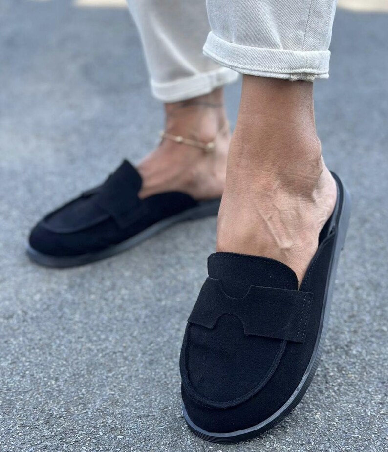 May include: A pair of black suede slip-on loafers featuring a rounded toe, a flat sole, and a wide decorative strap across the vamp with a cutout detail. These backless shoes have a minimalist design with a soft matte texture and tonal stitching.