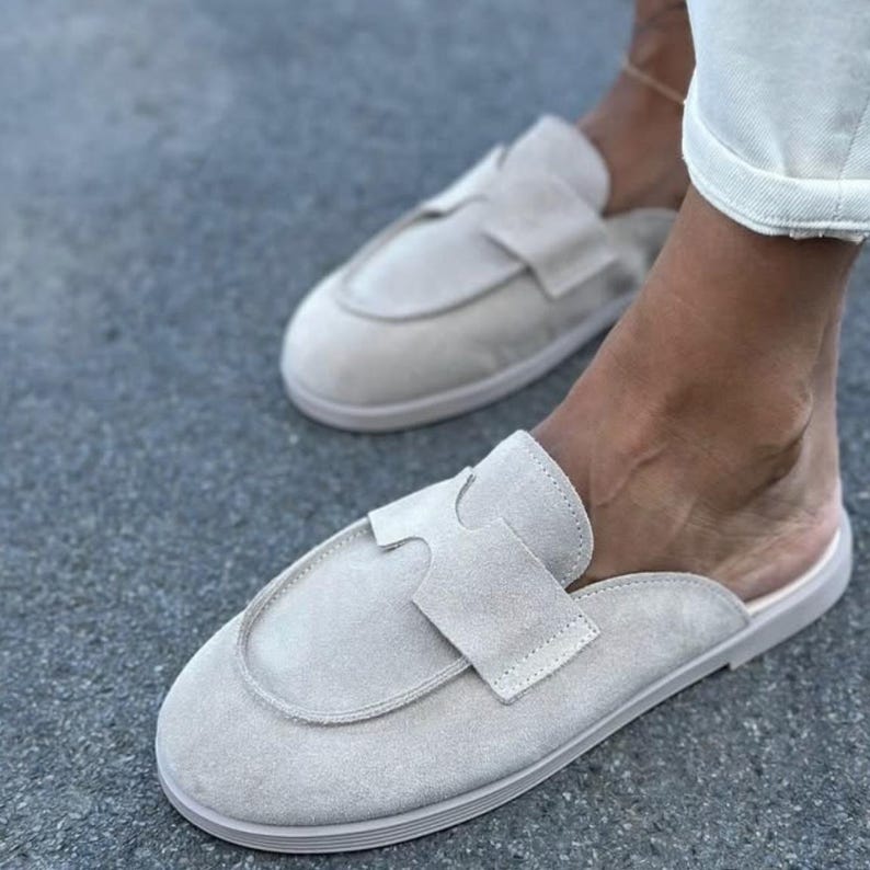 May include: Light beige suede slip-on shoes with a rounded toe and a decorative strap across the top. The shoes have a low profile and a neutral colour, suitable for casual wear. The soles are a matching light beige colour.