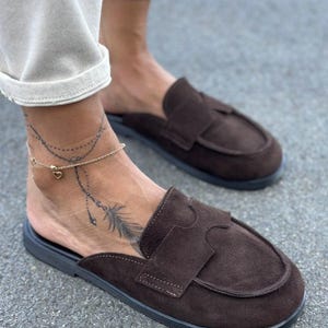 May include: A pair of dark brown suede backless loafers featuring a rounded toe, a wide strap across the vamp with a cutout detail, and a flat black sole. The shoes are slip-on style with an open heel design, showcasing a textured suede finish.