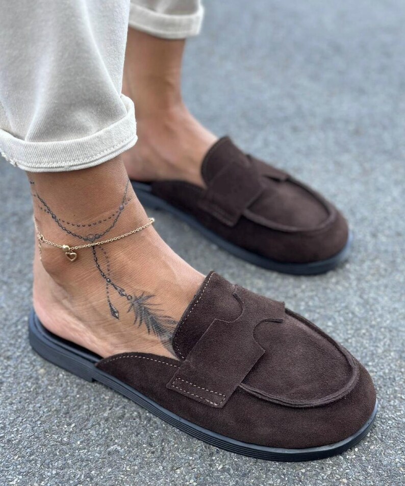 May include: A pair of dark brown suede backless loafers featuring a rounded toe, a wide strap across the vamp with a cutout detail, and a flat black sole. The shoes are slip-on style with an open heel design, showcasing a textured suede finish.