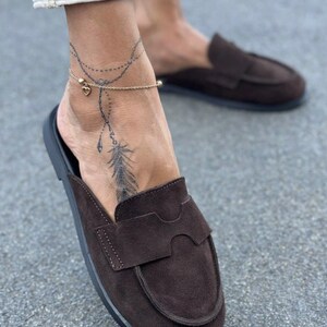 May include: A pair of dark brown suede slip-on loafers featuring a rounded toe, a stitched apron front, and a wide strap across the vamp. The shoes have a flat black sole and a soft, matte texture, providing a simple and functional footwear design.