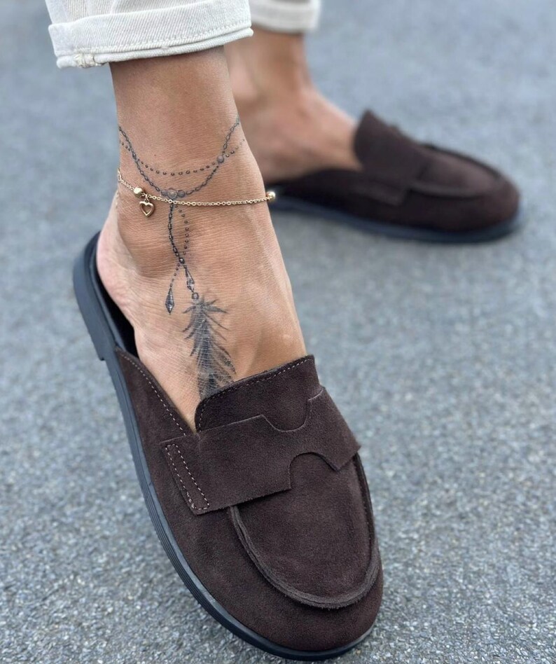 May include: A pair of dark brown suede slip-on loafers featuring a rounded toe, a stitched apron front, and a wide strap across the vamp. The shoes have a flat black sole and a soft, matte texture, providing a simple and functional footwear design.