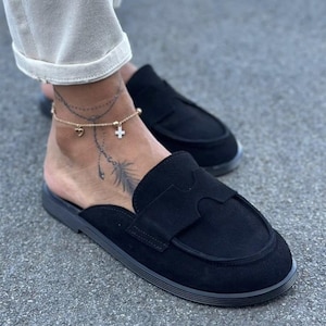 May include: A pair of black suede slip-on loafers featuring a rounded toe, a flat sole, and a decorative cutout strap across the vamp. The shoes have a low-profile design with an open back, showcasing a smooth texture and consistent black color.