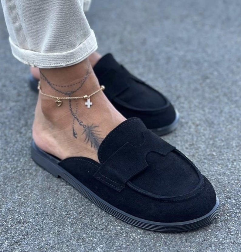 May include: A pair of black suede slip-on loafers featuring a rounded toe, a flat sole, and a decorative cutout strap across the vamp. The shoes have a low-profile design with an open back, showcasing a smooth texture and consistent black color.