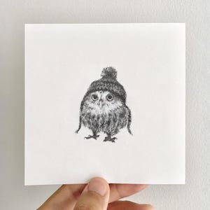 Può includere: Un disegno a carboncino in bianco e nero su carta bianca quadrata raffigurante un piccolo gufo che indossa un berretto lavorato a maglia con un pompon sulla sommità. Il gufo ha grandi occhi rotondi, piume dettagliate e artigli visibili.