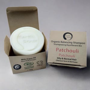 May include: A white, round shampoo bar with the text "Earth Sense" in a partially open cardboard box. The box next to it displays "Organic Balancing Shampoo Patchouli" and other text. The product is for oily and normal hair and contains Argan Oil.
