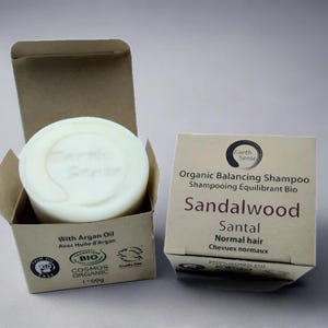 Può includere: Una saponetta di shampoo organico al legno di sandalo in una scatola di cartone. La saponetta è rotonda e bianco sporco, con le parole "Earth Sense" in rilievo. La scatola mostra anche le parole "Capelli normali" e "60g".