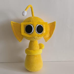 May include: A plush yellow creature with large black eyes, wearing yellow square-framed glasses. It has large, pointed ears and a curved antenna with a white ball at the end. A yellow ring encircles the base of the neck. The plush toy is on a white background.