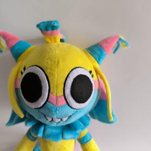 K&ouml;nnte beinhalten: Ein Pl&uuml;schtier mit einem leuchtend gelben Kopf, gro&szlig;en schwarzen runden Augen, einer hellblauen Schnauze und einer rosa Nase. Die Figur hat spitze Ohren mit rosa und blauen Akzenten, gelbe Haarstr&auml;hnen und einen blauen R&uuml;schenkragen.