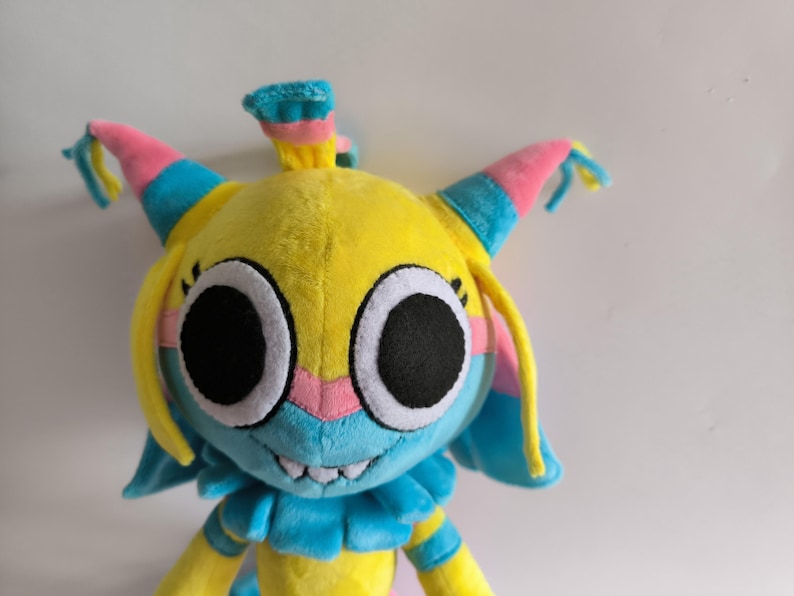 K&ouml;nnte beinhalten: Ein Pl&uuml;schtier mit einem leuchtend gelben Kopf, gro&szlig;en schwarzen runden Augen, einer hellblauen Schnauze und einer rosa Nase. Die Figur hat spitze Ohren mit rosa und blauen Akzenten, gelbe Haarstr&auml;hnen und einen blauen R&uuml;schenkragen.