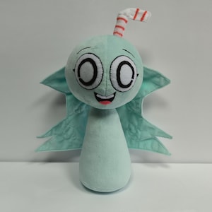 Op de afbeelding: Een pluche pop in de vorm van een stripfiguur met een lichtblauwe kleur, grote ogen en een lachende mond. Het heeft blauwe vleugels en een gestreepte antenne. De pop is gemaakt van zachte stof.