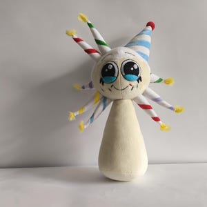 Könnte beinhalten: Ein Plüschtier mit einem skurrilen Design. Es hat einen cremefarbenen Körper und einen Kopf mit großen, ausdrucksstarken Augen und einem lächelnden Mund. Der Kopf ist mit gestreiften, kegelförmigen Anhängseln und einer Partyhütchen verziert. Das Spielzeug ist etwa 20 cm groß.
