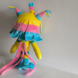 K&ouml;nnte beinhalten: Ein Pl&uuml;schtier in den Farben Gelb, Pink und Blau. Die Kreatur hat einen abgerundeten Kopf mit spitzen, bunten Ohren, einen ger&uuml;schten Kragen und lange, d&uuml;nne pinke Anh&auml;ngsel, die vom R&uuml;cken herabh&auml;ngen. Es steht auf einer blauen Basis.