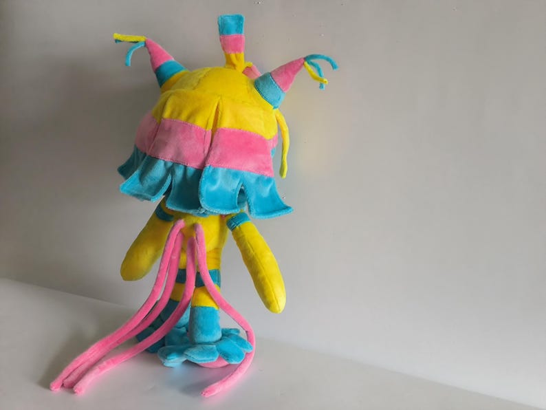 K&ouml;nnte beinhalten: Ein Pl&uuml;schtier in den Farben Gelb, Pink und Blau. Die Kreatur hat einen abgerundeten Kopf mit spitzen, bunten Ohren, einen ger&uuml;schten Kragen und lange, d&uuml;nne pinke Anh&auml;ngsel, die vom R&uuml;cken herabh&auml;ngen. Es steht auf einer blauen Basis.