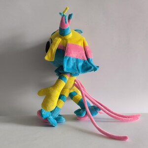 K&ouml;nnte beinhalten: Ein Pl&uuml;schtier-Charakter mit gelbem Kopf, blauen und rosa Akzenten, gro&szlig;en schwarzen Augen und hohen spitzen Ohren. Der K&ouml;rper hat gelb-blau gestreifte Gliedma&szlig;en, rosa R&uuml;schenmanschetten und zwei lange, d&uuml;nne rosa Schw&auml;nze am hinteren Teil.