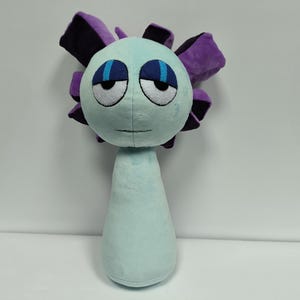 Puede incluir: Un peluche con forma de flor estilizada, con cuerpo y cabeza azul claro. La flor tiene ojos grandes y somnolientos con detalles azules y pétalos morados y negros. El juguete está diseñado para ser suave y tierno.