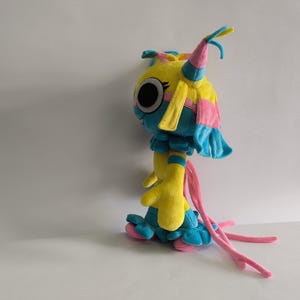 K&ouml;nnte beinhalten: Ein Pl&uuml;schtier in Form einer gelb-blauen Kreatur mit gro&szlig;en schwarzen Augen, rosa Akzenten und langen, herabh&auml;ngenden rosa B&auml;ndern. Die Figur hat einen runden Kopf mit spitzen Ohren, einen ger&uuml;schten Kragen und einen rockartigen Saum.