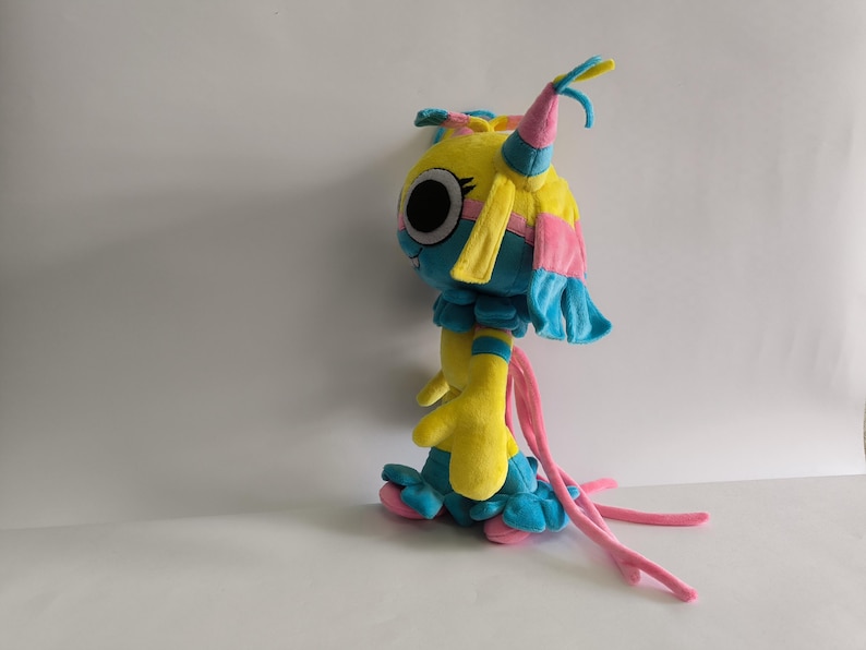 K&ouml;nnte beinhalten: Ein Pl&uuml;schtier in Form einer gelb-blauen Kreatur mit gro&szlig;en schwarzen Augen, rosa Akzenten und langen, herabh&auml;ngenden rosa B&auml;ndern. Die Figur hat einen runden Kopf mit spitzen Ohren, einen ger&uuml;schten Kragen und einen rockartigen Saum.