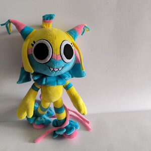 K&ouml;nnte beinhalten: Ein Pl&uuml;schtier in den Farben Gelb, Blau und Pink. Die Figur hat gro&szlig;e schwarze Augen, ein breites Grinsen mit sichtbaren Z&auml;hnen und einen ger&uuml;schten Kragen. Es verf&uuml;gt &uuml;ber lange, herabh&auml;ngende Gliedma&szlig;en mit rosa B&auml;ndern und eine spitze Kopfbedeckung.