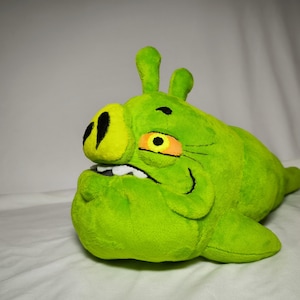Op de afbeelding: Een groene pluche Angry Birds varkensfiguur met een grote snuit, kleine oren en een boze uitdrukking. Het varken heeft een gele neus en oog, en witte tanden. Het pluche speelgoed ligt op een witte ondergrond.