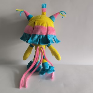 K&ouml;nnte beinhalten: Ein Pl&uuml;schtier in den Farben Gelb, Pink und Blau. Die Kreatur hat einen gro&szlig;en, abgerundeten Kopf mit mehreren spitzen, hornartigen Forts&auml;tzen, lange herabh&auml;ngende pinke Tentakel und blaue, bl&uuml;tenblattartige F&uuml;&szlig;e. Das Stofftier steht aufrecht.