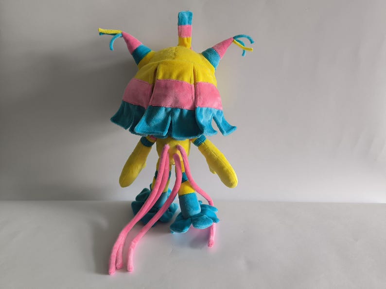 K&ouml;nnte beinhalten: Ein Pl&uuml;schtier in den Farben Gelb, Pink und Blau. Die Kreatur hat einen gro&szlig;en, abgerundeten Kopf mit mehreren spitzen, hornartigen Forts&auml;tzen, lange herabh&auml;ngende pinke Tentakel und blaue, bl&uuml;tenblattartige F&uuml;&szlig;e. Das Stofftier steht aufrecht.