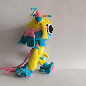 K&ouml;nnte beinhalten: Ein Pl&uuml;schtier mit gelbem Kopf und K&ouml;rper sowie gro&szlig;en, schwarzen, kreisf&ouml;rmigen Augen. Die Figur hat blaue und rosa Akzente, darunter lange, haar&auml;hnliche Str&auml;hnen, eine spitz zulaufende Kopfbedeckung sowie blaue R&uuml;schen an Hand- und Fu&szlig;gelenken.