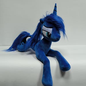 Könnte beinhalten: Ein Plüschtier in Form eines blauen Ponys mit langer, dunkelblauer Mähne und Schweif, einem einzelnen Horn auf der Stirn und großen, gestickten Augen mit hellblauen Iris. Die Figur ist in einer liegenden Position mit ausgestreckten Vorderbeinen.